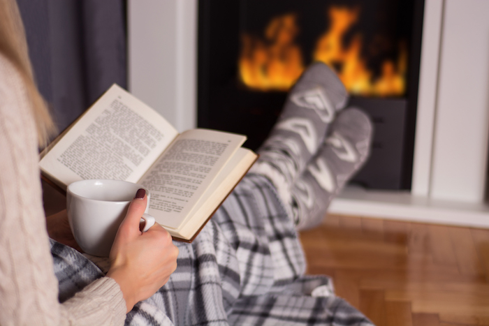 blonde female sitting by the fire with a book and cup of coffee